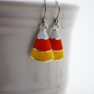 A pair of silver plated enamel candy corn earrings with orange, yellow, and white coloring, displayed over a white ceramic dish.